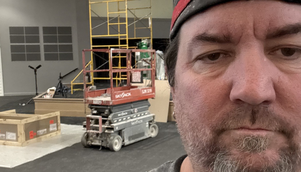 Close-up selfie of a man on a construction site, with a red scissor lift and scaffolding behind him.