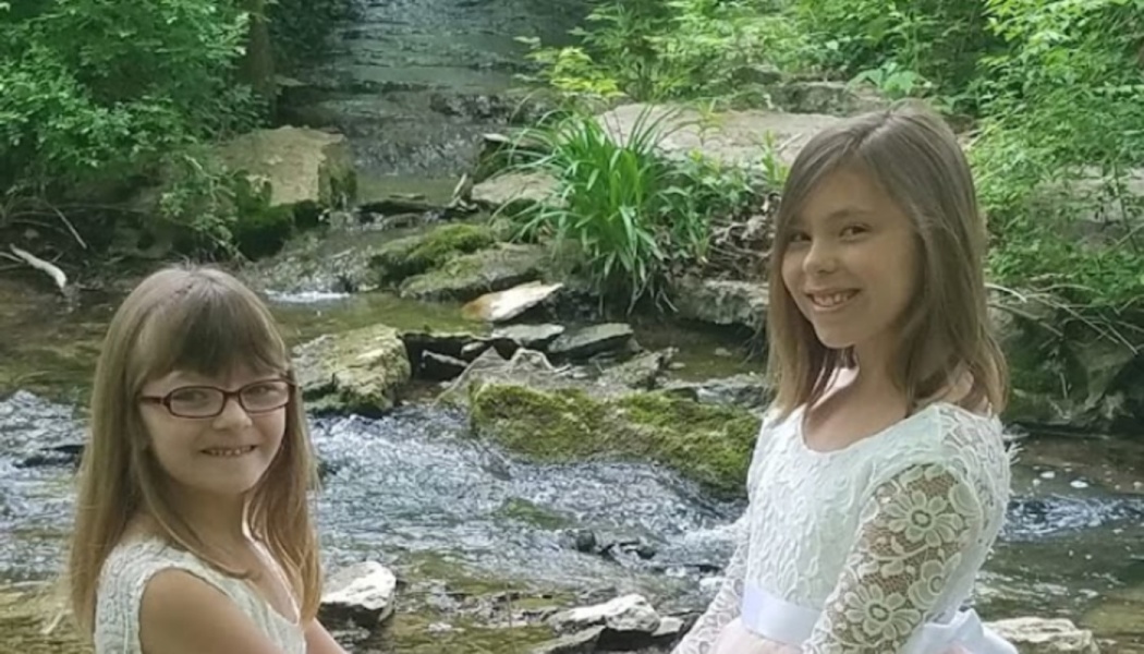 Two smiling young girls in white lace dresses pose outdoors in front of a rocky forest stream.