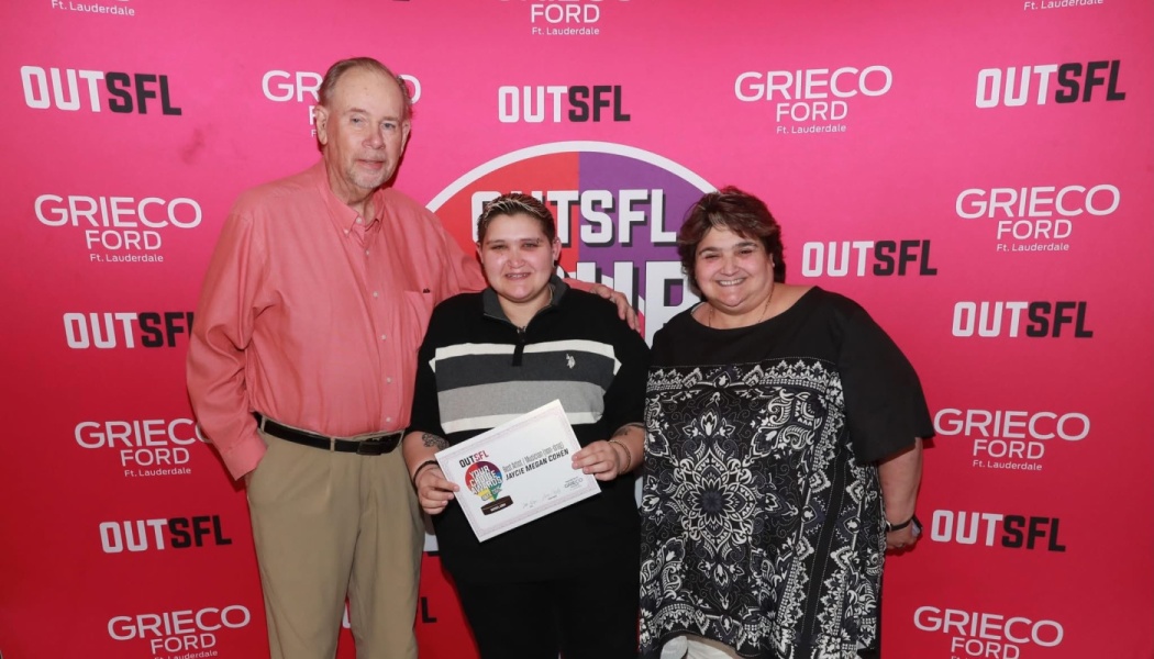Three people pose in front of a red backdrop as the center person holds an OUTSFL award certificate.