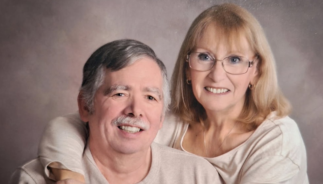 A smiling older couple, with a blonde woman in glasses embracing a man with grey hair and a mustache.