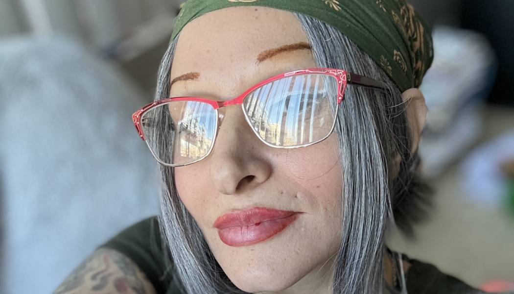 Close-up portrait of a woman with straight grey hair, red cat-eye glasses, red lipstick, and a green bandana.
