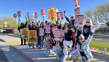 Striking educators holding red and yellow signs that read "On Strike for Our Students" on a sunny sidewalk.