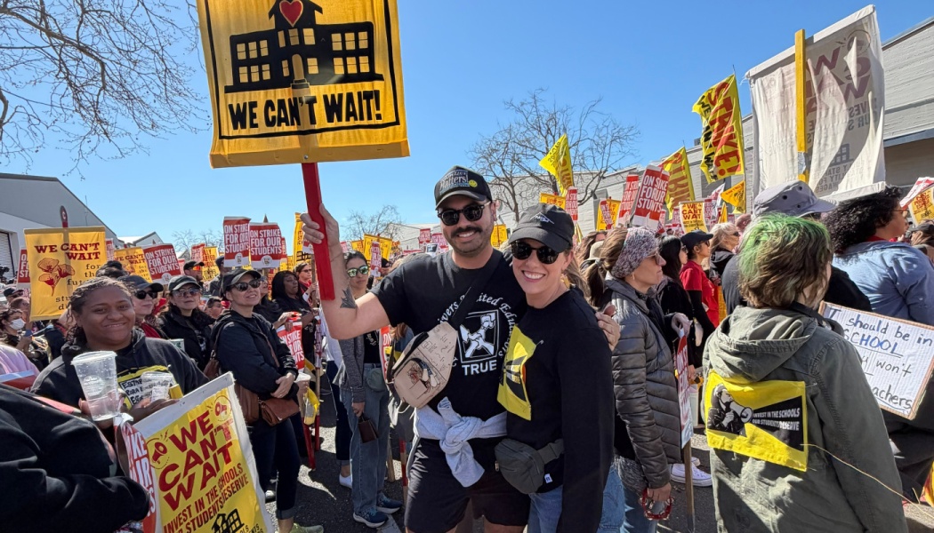 Two smiling demonstrators stand in a crowd of education protesters holding yellow "WE CAN'T WAIT!" signs.