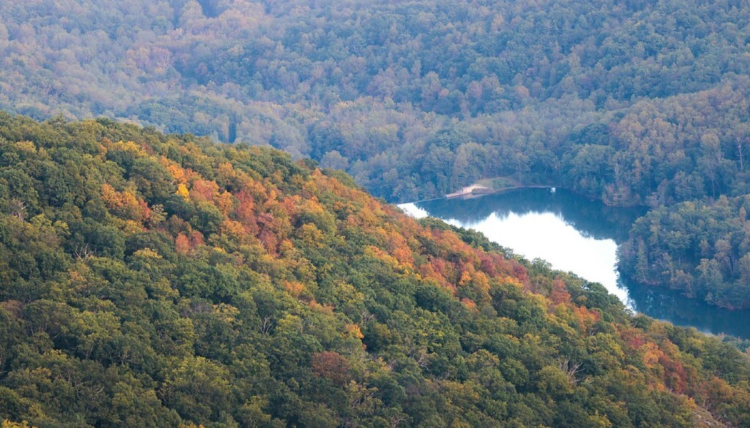 Aerial view of autumn foliage covering forested hillsides surrounding a serene lake in a valley.