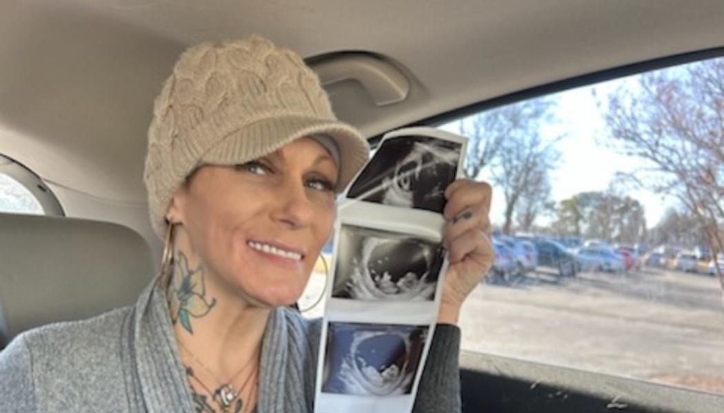 A smiling woman in a knit hat holds up a strip of baby ultrasound scan photos while sitting inside a car.