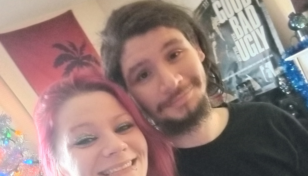 A smiling woman with bright pink hair and a bearded man posing closely together for an indoor selfie.