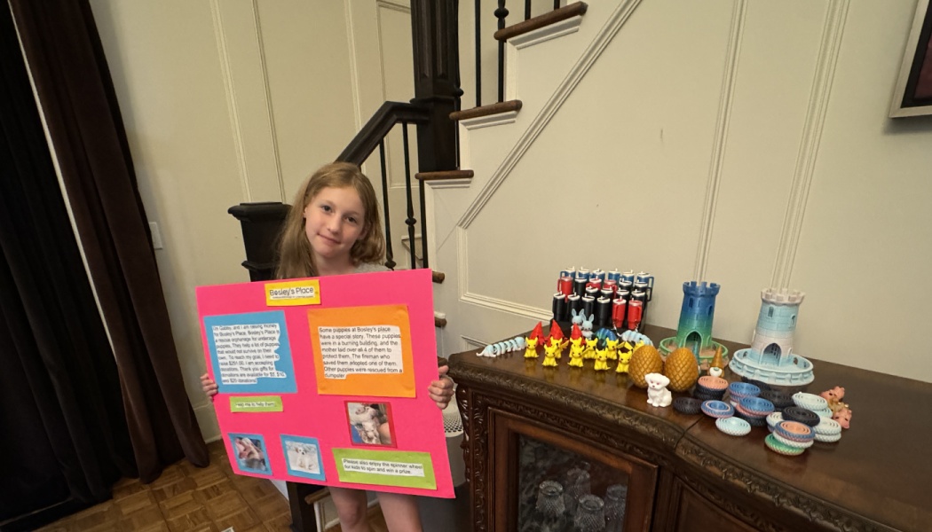 A young girl holds a pink poster board for a puppy rescue fundraiser next to a table of 3D-printed toys.