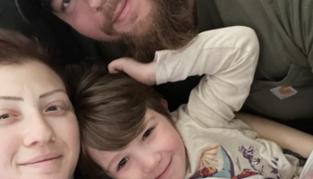 A close-up selfie of a mother, bearded father, and young child lying close together and smiling.