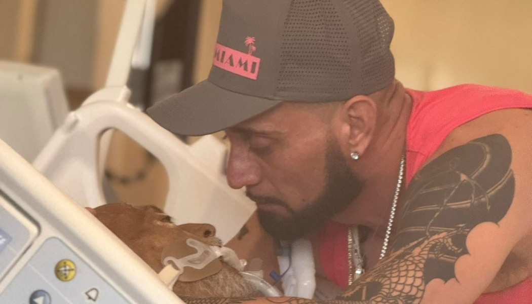 A tattooed man in a baseball cap leaning close to a loved one in a hospital bed with medical tubes.