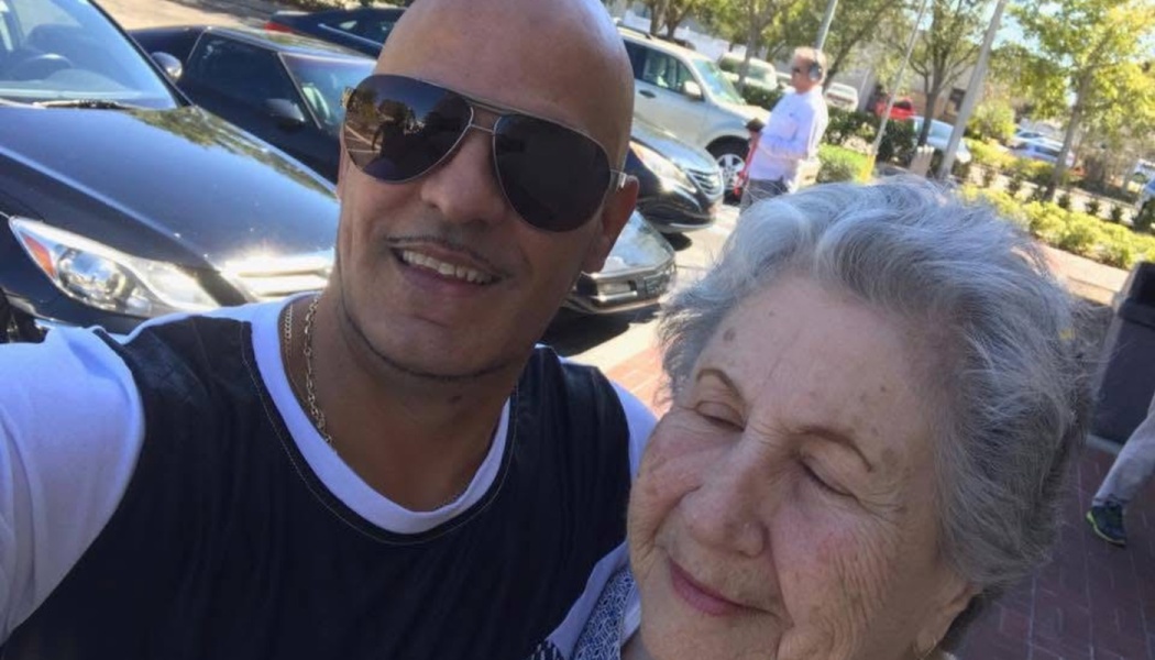 A smiling bald man wearing sunglasses takes a selfie with an elderly woman with short gray hair outdoors.