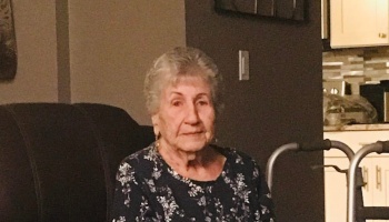 An elderly woman with short grey hair in a floral shirt sitting indoors next to a walker.