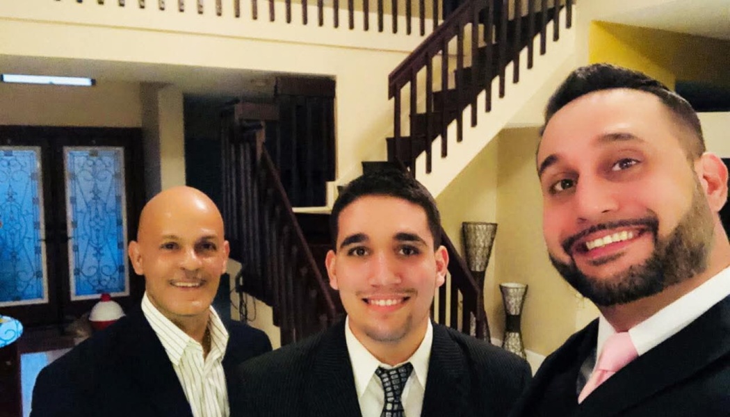 Three smiling men dressed in suits pose for a selfie inside a home with a staircase behind them.