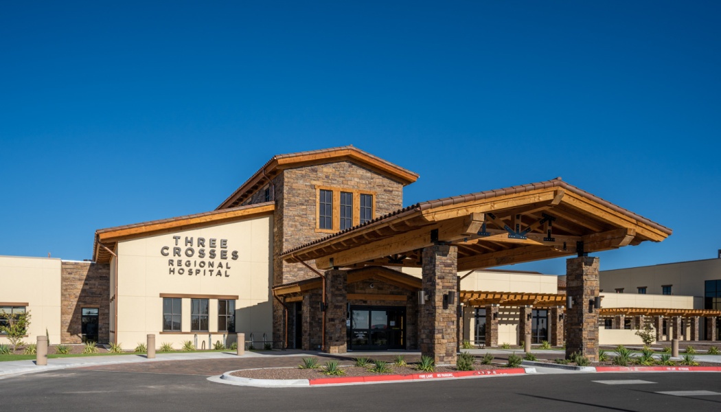 Exterior of Three Crosses Regional Hospital with stone accents and a wooden covered entryway under a clear blue sky.