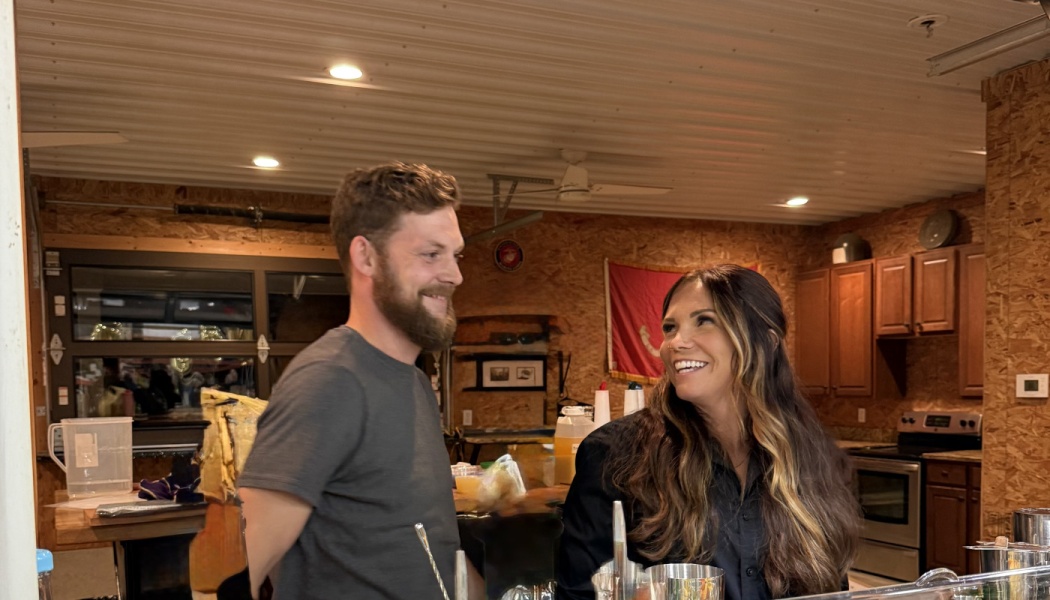 A bearded man and a long-haired woman smile happily at each other behind a rustic wooden bar.