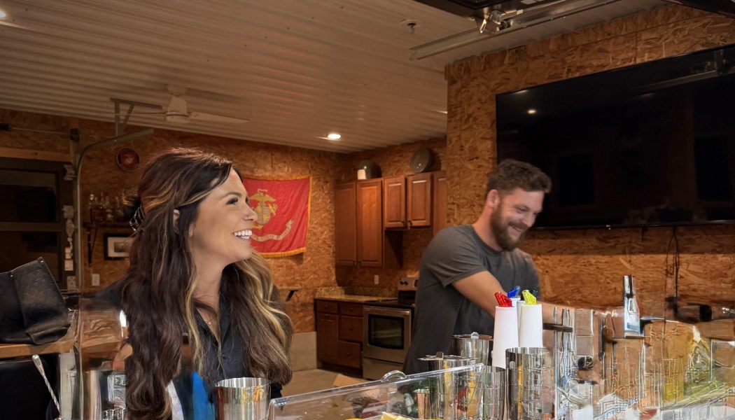 A smiling woman and bearded man behind a rustic bar with cocktail shakers in the foreground.