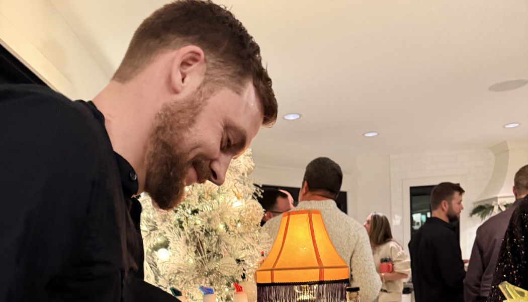 A smiling bearded man looking down at a festive holiday party with people mingling near a decorated tree.