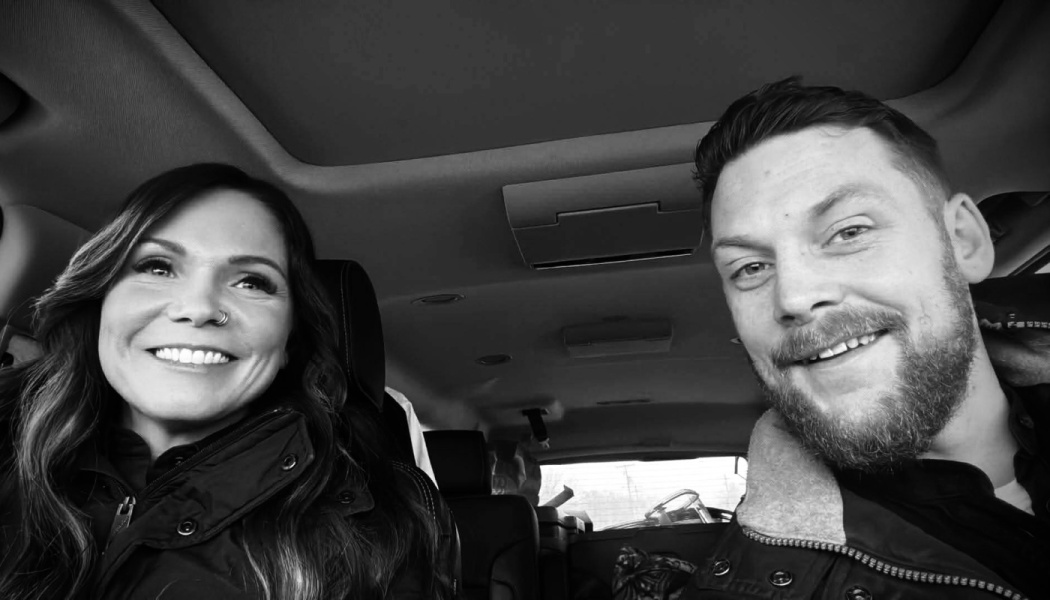 Black and white selfie of a smiling woman and bearded man sitting inside a car.
