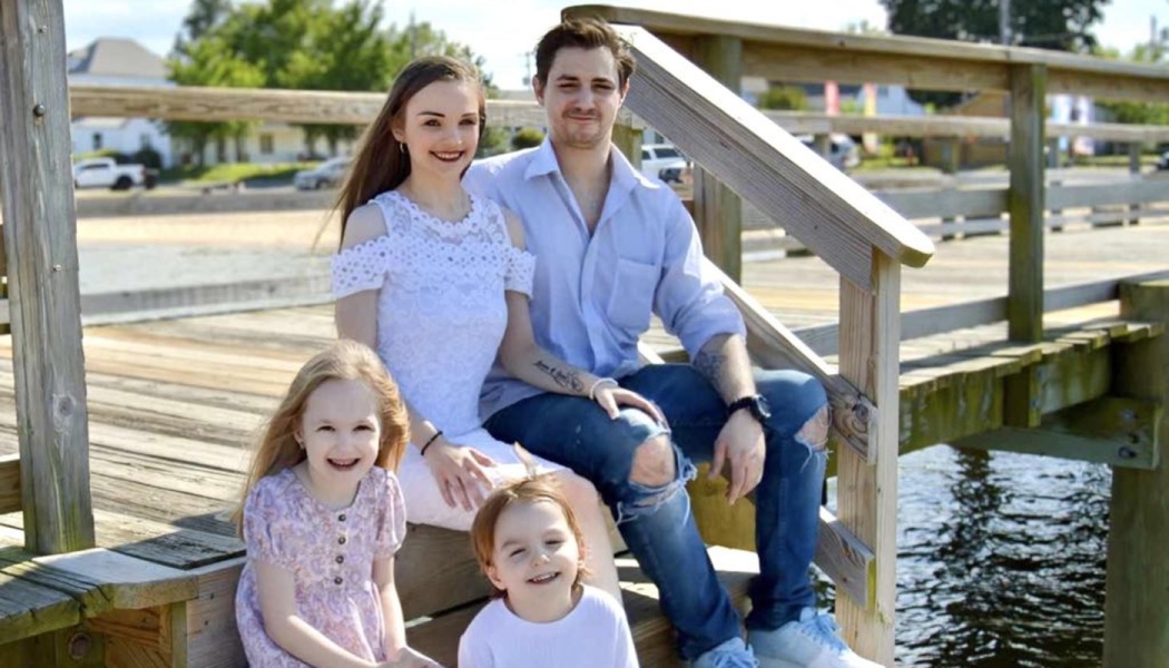 A smiling family of four with two young children sitting on the wooden stairs of a waterfront dock.