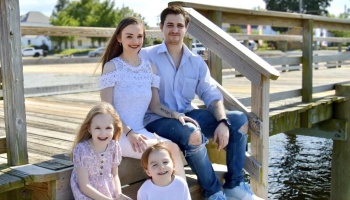 A smiling family of four with two young children sitting on the wooden stairs of a waterfront dock.