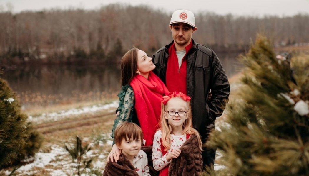 A family of four with two young children posing together outdoors near pine trees and a lake in winter.