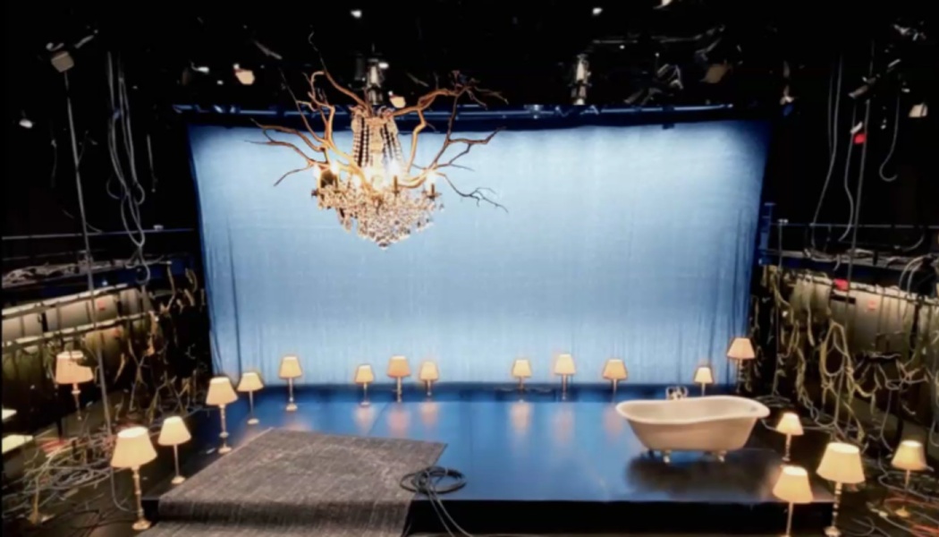 A surreal theater stage with a blue backdrop, white bathtub, floor lamps, and a chandelier woven with branches.