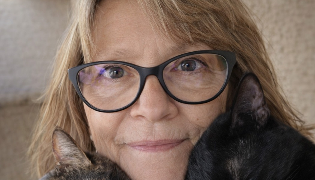 A smiling woman wearing glasses cuddles a black cat and a tabby cat close to her face.