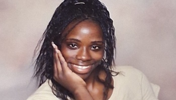 A smiling Black woman with dark braided hair resting her cheek on her hand.