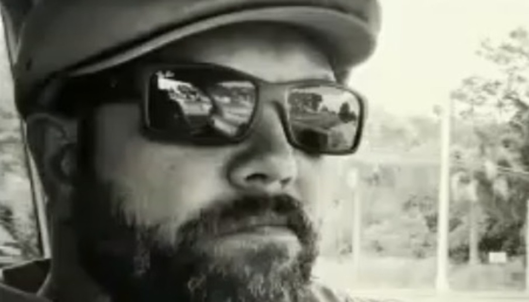 Black and white close-up selfie of a bearded man wearing sunglasses and a cap.