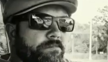 Black and white close-up selfie of a bearded man wearing sunglasses and a cap.