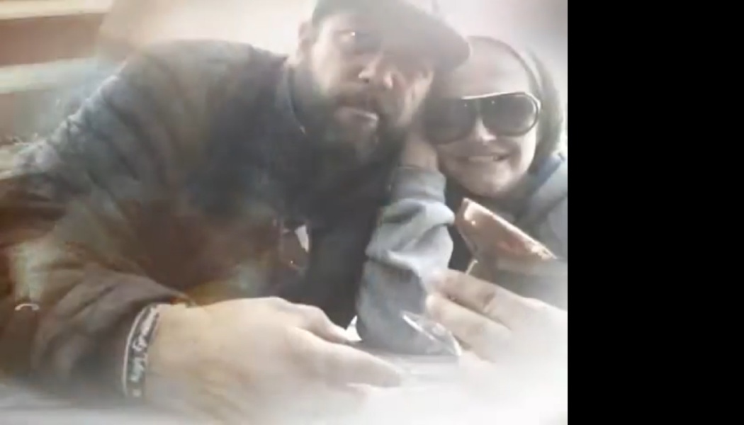 A bearded man and a smiling woman in large sunglasses pose closely together.