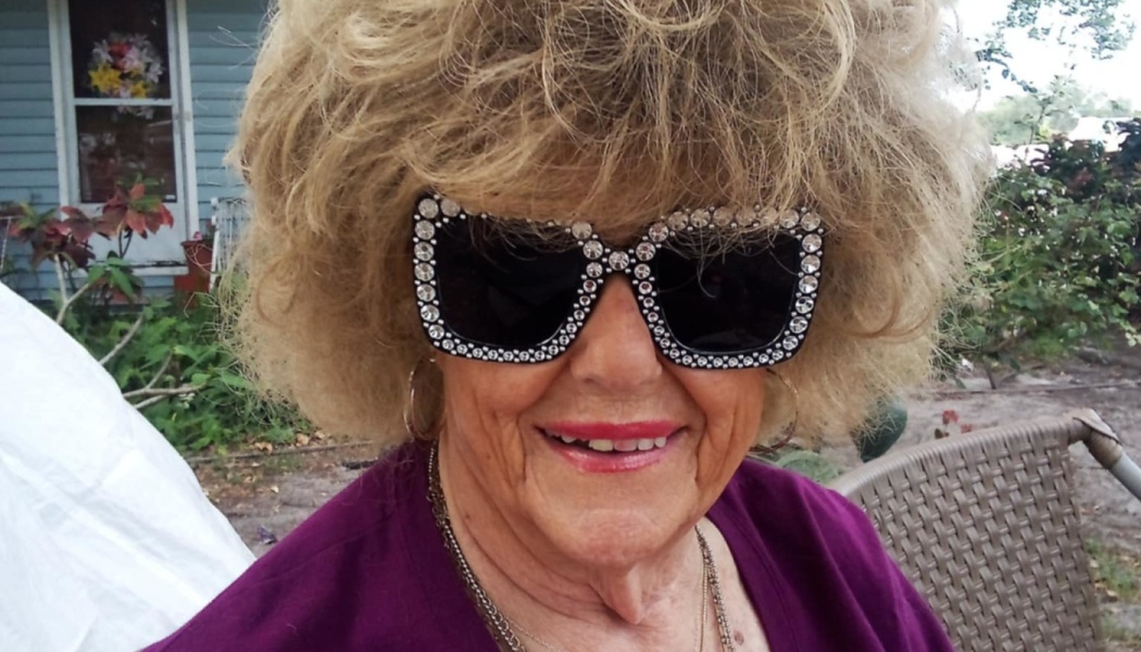 Smiling woman with big blonde hair wearing oversized rhinestone sunglasses and a purple top.