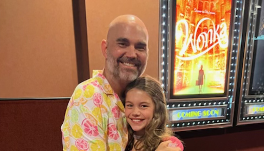 A smiling man in a citrus-print shirt hugging a young girl at a movie theater next to a Wonka poster.