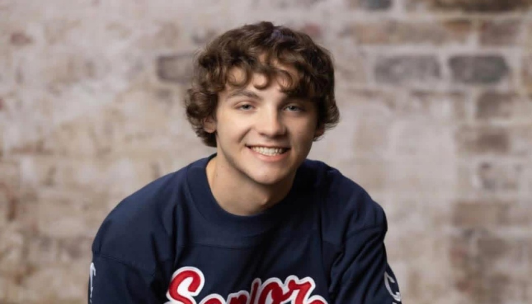 A smiling teenage boy with curly brown hair wearing a dark blue shirt in front of a brick wall.