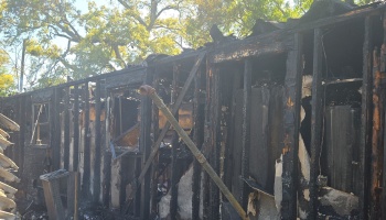 Extensively charred exterior wall and exposed wooden frame of a house destroyed by fire.
