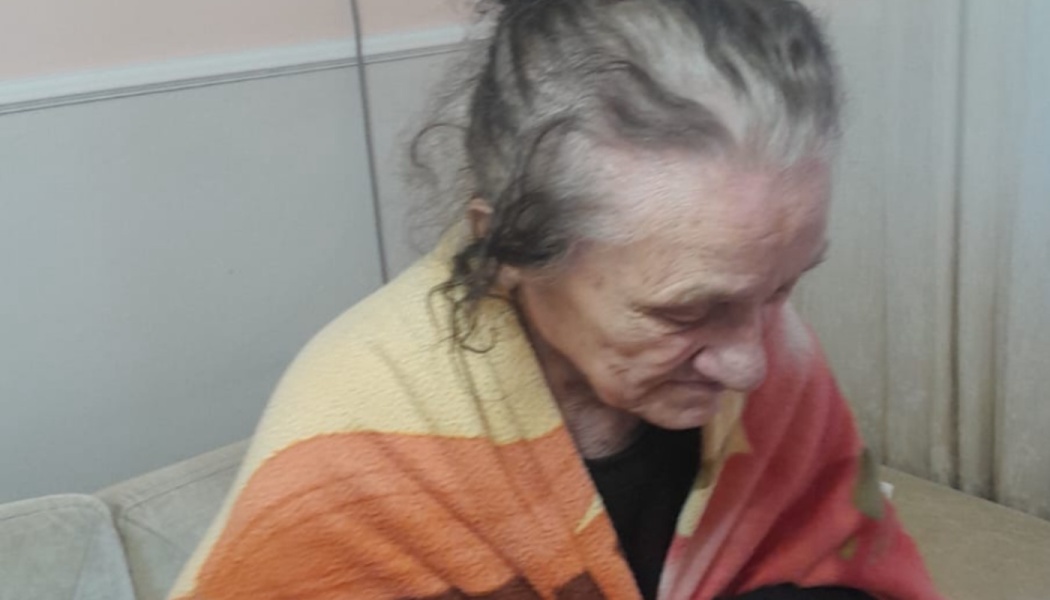 An elderly woman with grey hair wrapped in a colorful blanket, looking downwards.