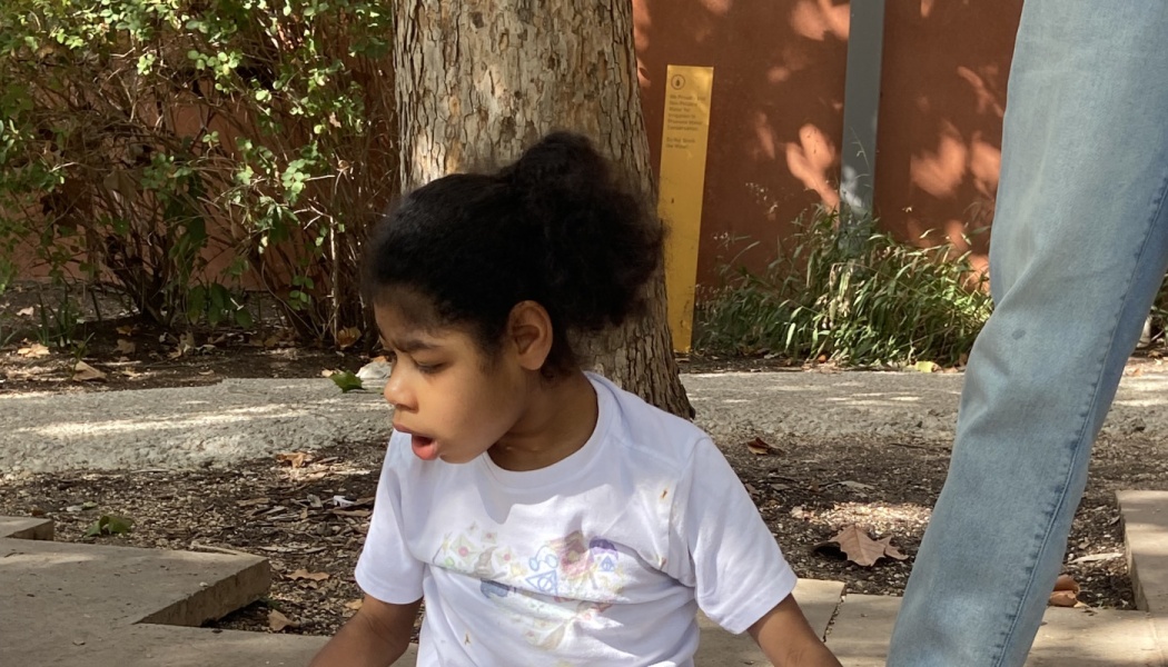 A young girl with dark curly hair wearing a white t-shirt sits outdoors near a tree trunk.
