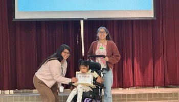 A young girl in a wheelchair holds an attendance certificate on stage with two smiling women.