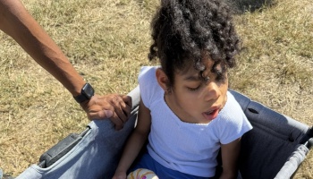 Young girl with curly hair sitting in a gray wagon while an adult's hand rests on the edge.