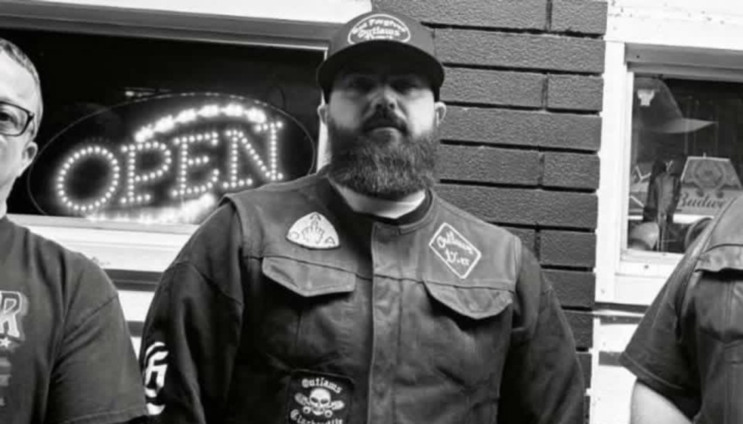 A bearded man in a cap and patched vest stands in front of a shop with a glowing OPEN sign.