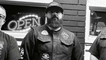 A bearded man in a cap and patched vest stands in front of a shop with a glowing OPEN sign.