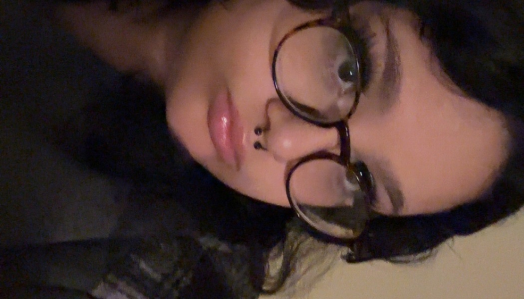 Close-up portrait of a person with dark hair, round glasses, and a septum piercing.