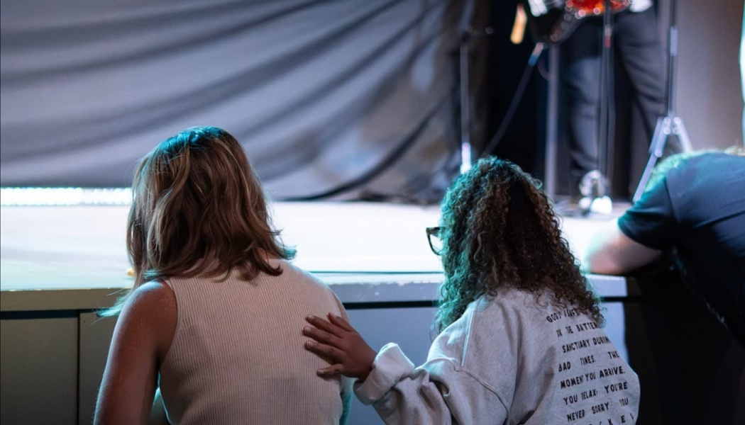 A woman with curly hair places a comforting hand on another woman's back near a stage.