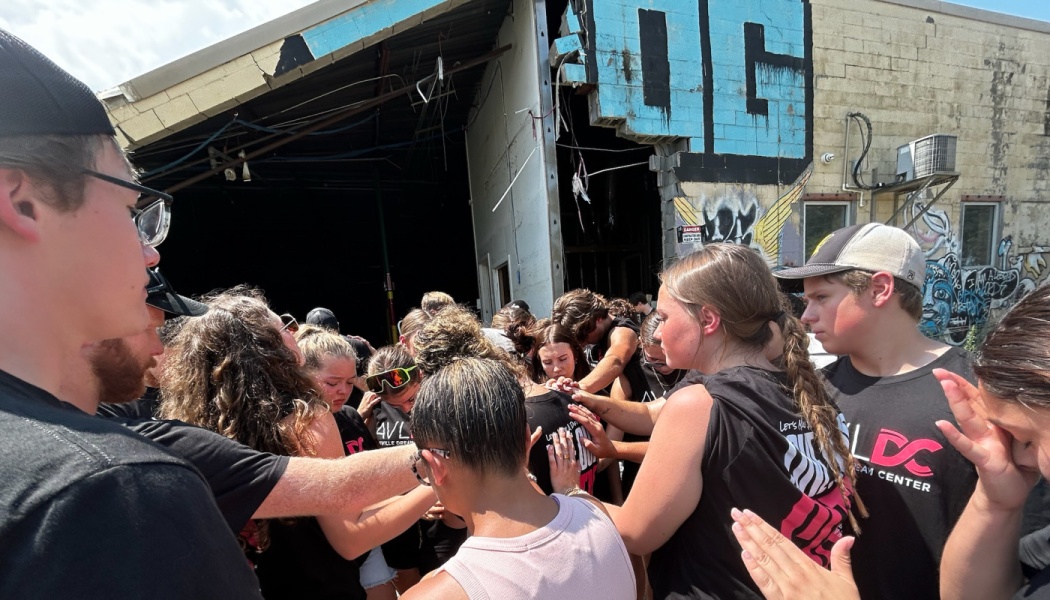 A group of young people huddle together in prayer outside a damaged building.
