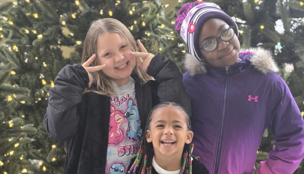 Three smiling young girls pose in winter coats in front of a lit Christmas tree.