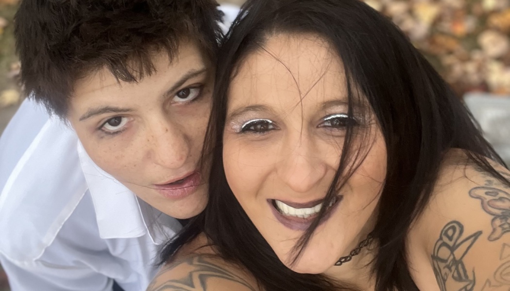 Close-up selfie of a couple outdoors, one smiling with long dark hair and shoulder tattoos, the other with short hair.