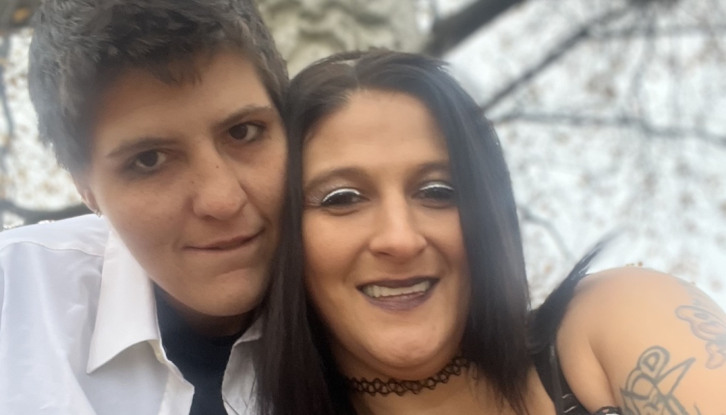 A smiling couple posing close together outdoors, one with short hair and the other with long dark hair.