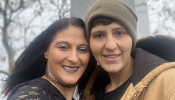 A woman with long dark hair and a choker smiling closely with a person wearing a beanie and tan jacket.