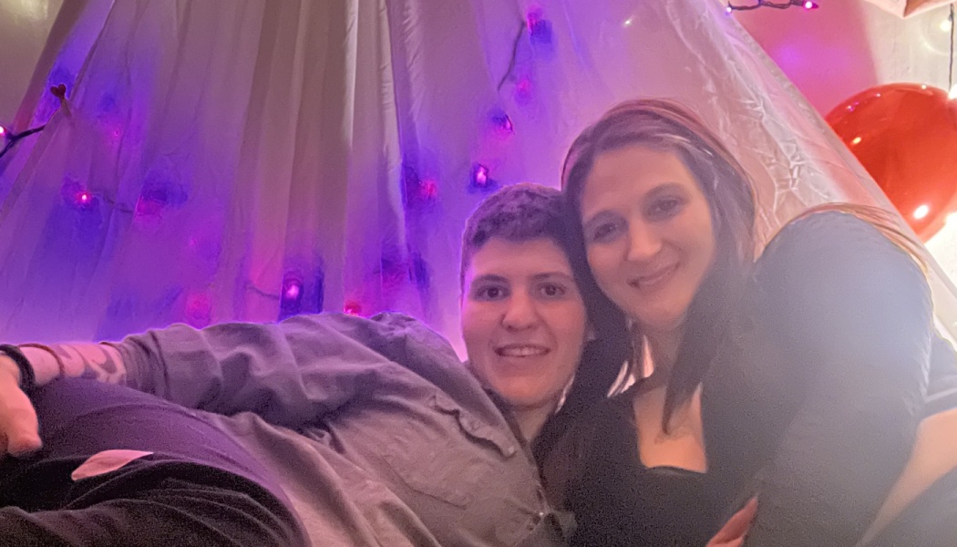 A smiling couple posing closely together under a cozy canopy illuminated with purple fairy lights.
