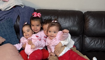 Four young siblings wearing matching pink and red outfits cuddle happily together on a dark leather sofa.