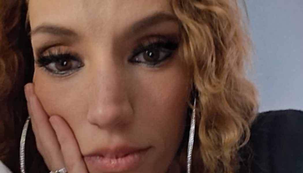 Close-up portrait of a woman with curly blonde hair resting her cheek on her hand, wearing dark eye makeup.